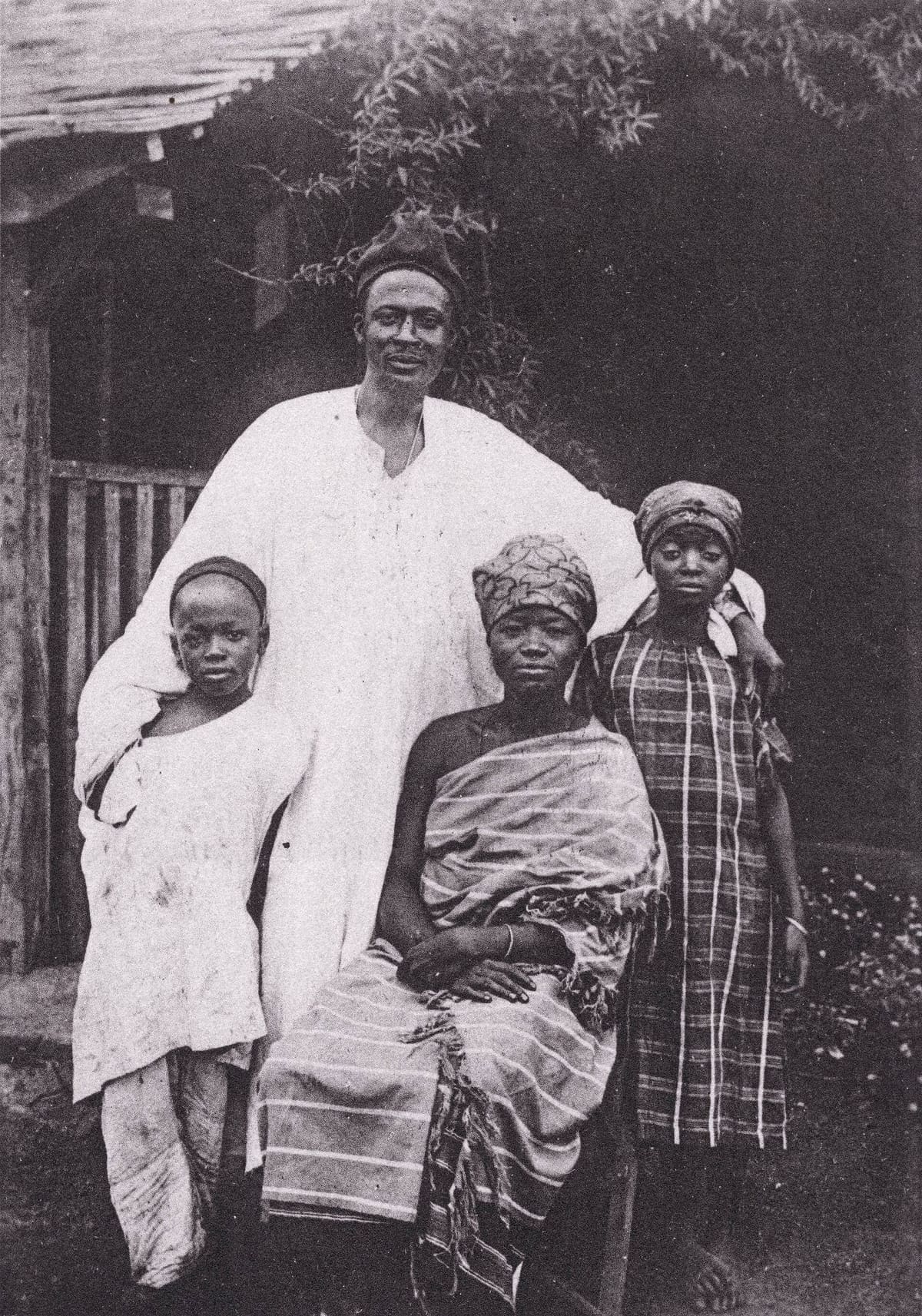 A Cameroonian family of four posed outside a house in a sepia print: a man standing in a long white gown, a woman seated in front of him wrapped in striped cloth, a child leaning against his side and an older girl standing behind in a checked dress.
