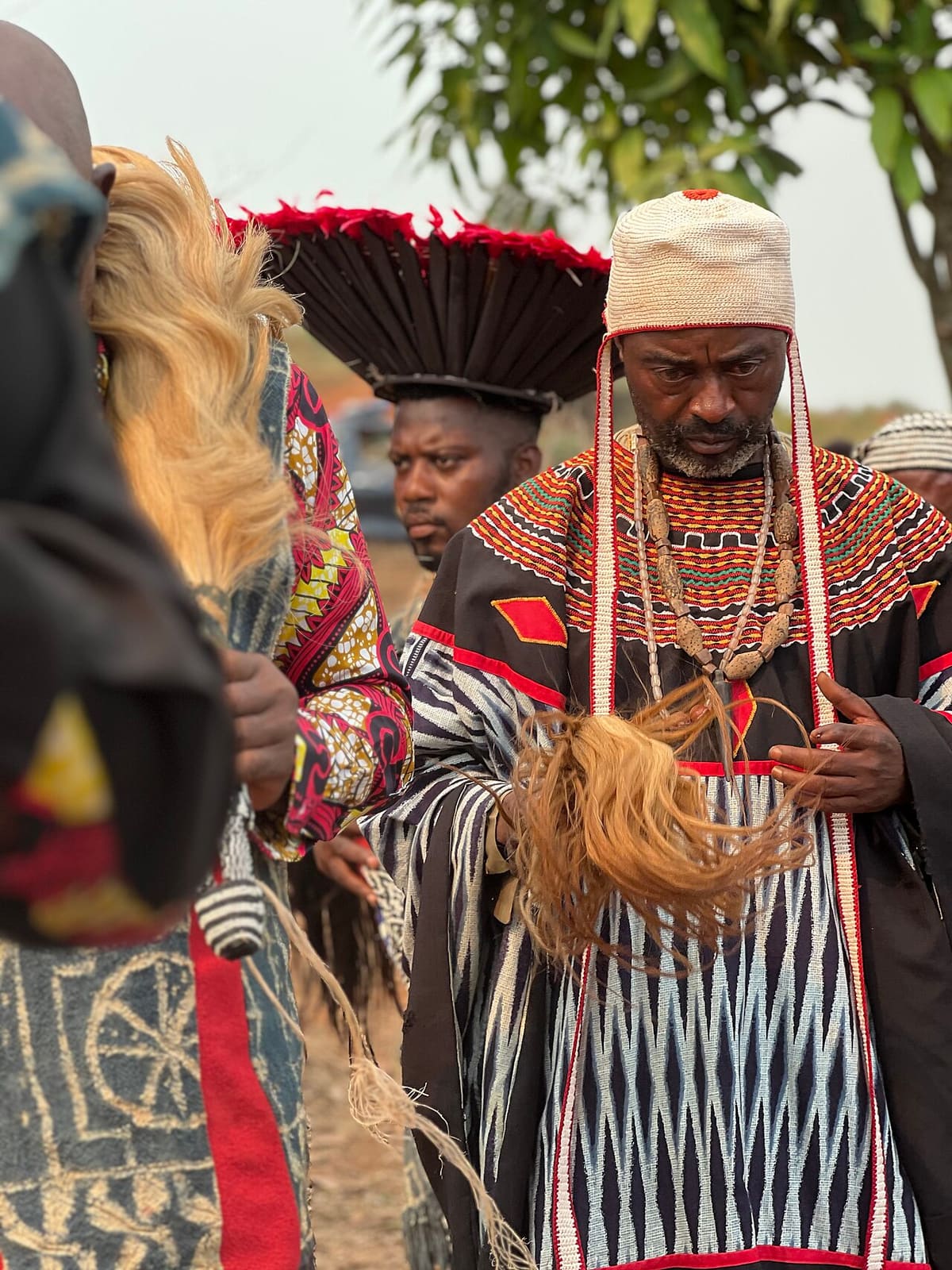 An older man in embroidered Grassfields robes and a woven cap, wearing beaded regalia, standing among other men in ceremonial dress at an outdoor gathering.