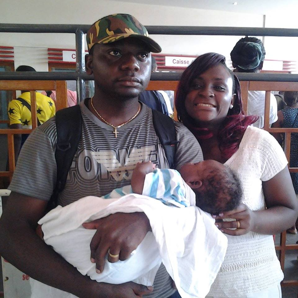A man and a woman standing close together indoors, the man holding a sleeping newborn wrapped in a white blanket, the woman resting her hand on the baby.