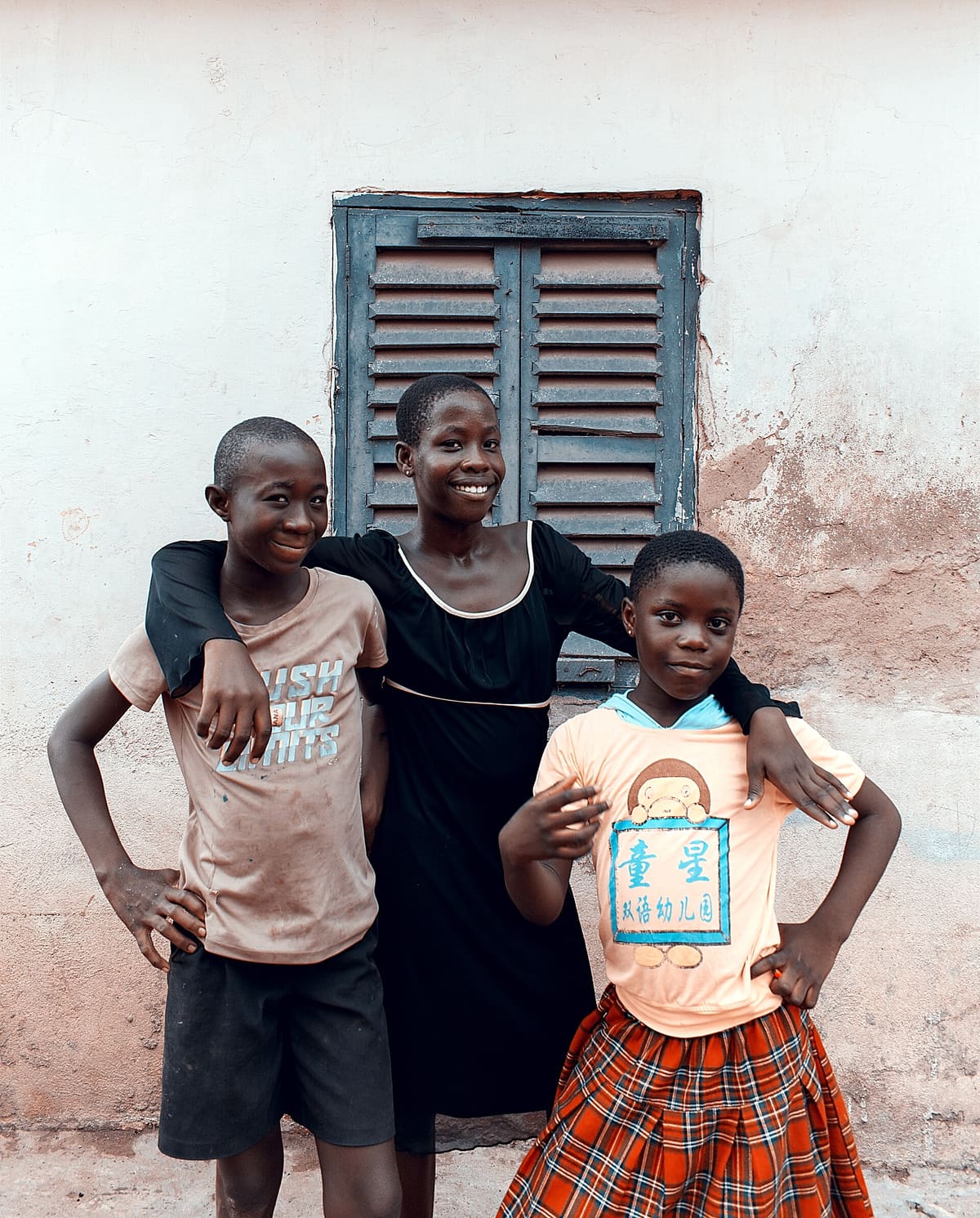 Three young siblings standing arm in arm against a pale wall beside a shuttered window, two of them smiling at the camera.