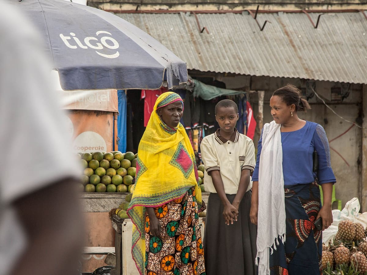 An older woman in a bright yellow headscarf, a girl in a cream shirt and a younger woman in blue standing together in front of a fruit stall under a market umbrella.
